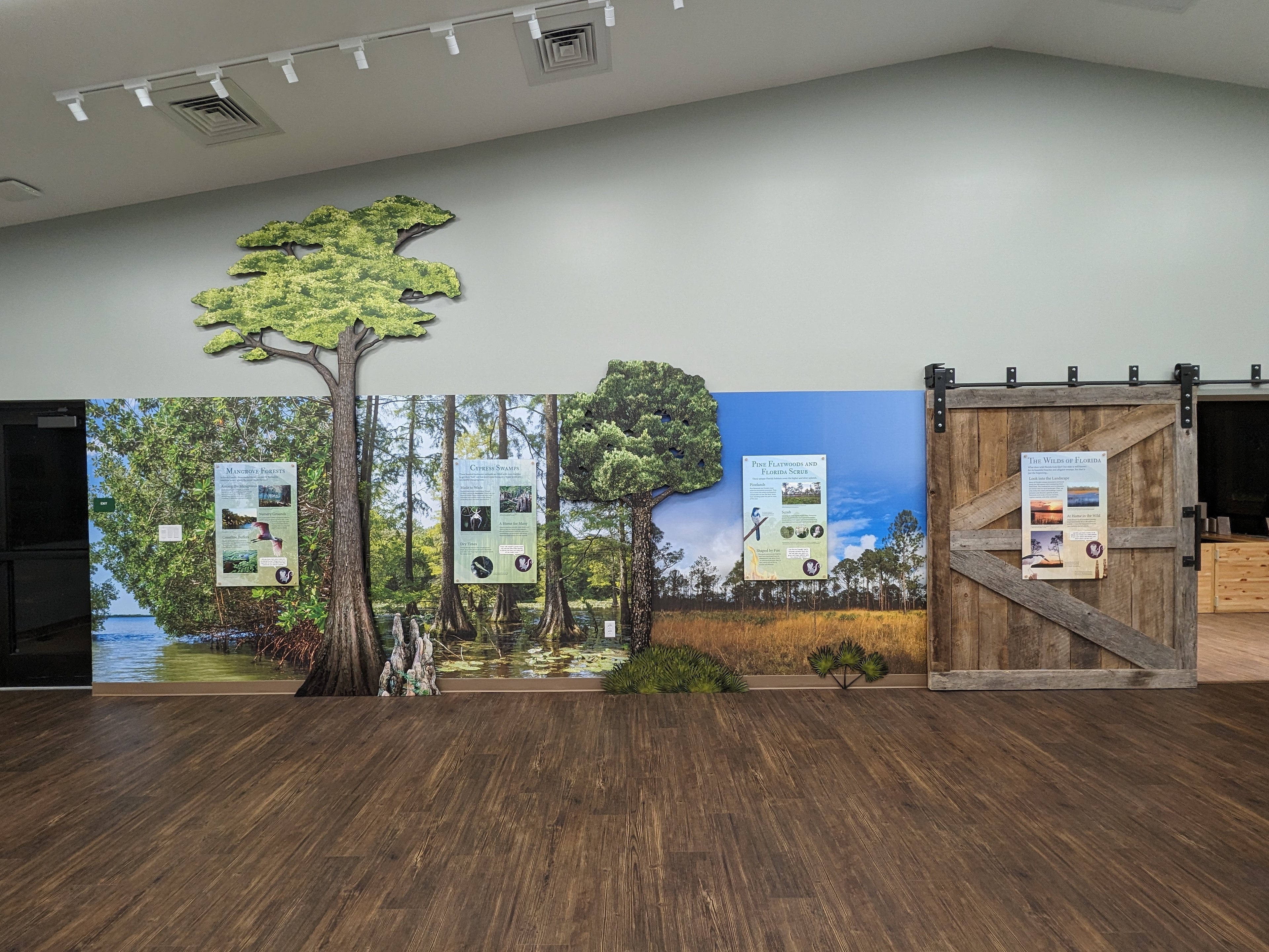 Exhibition space with large wall mural of a forest scene and wooden door.