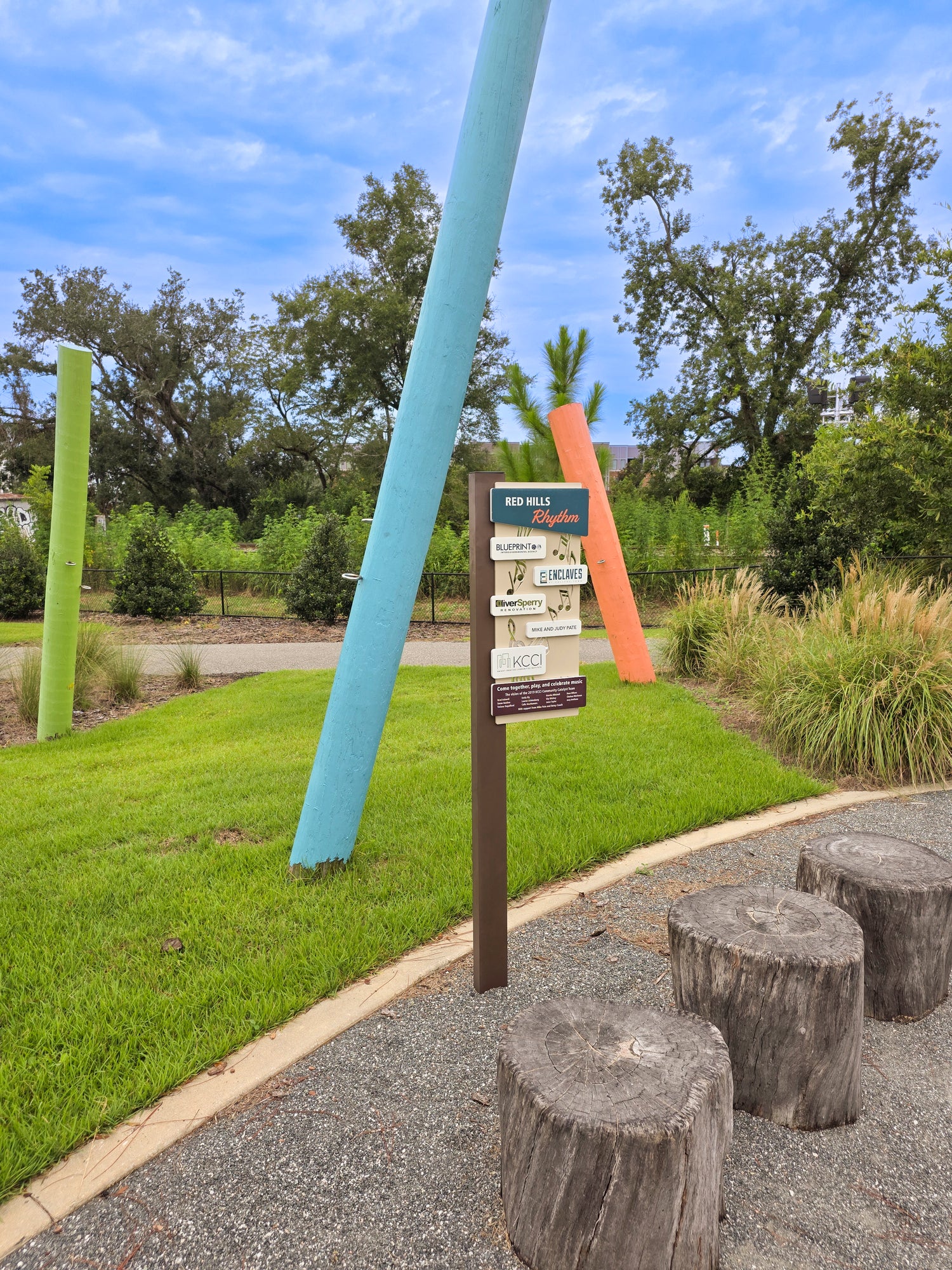 An outdoor park scene with a gravel path, tree-stump stools, a grassy lawn, and colorful sculptural poles. A small signpost is visible near the center, with trees and blue sky in the background.