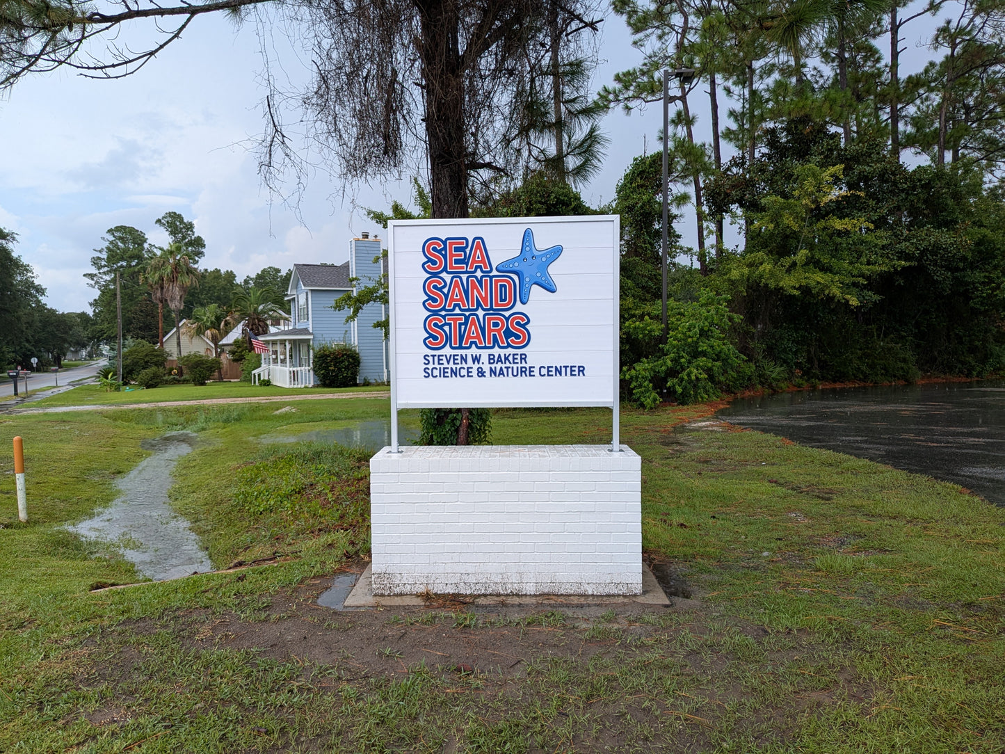 Sign for Sea Sand Stars Nature Center in a grassy area with trees and a house in the background.