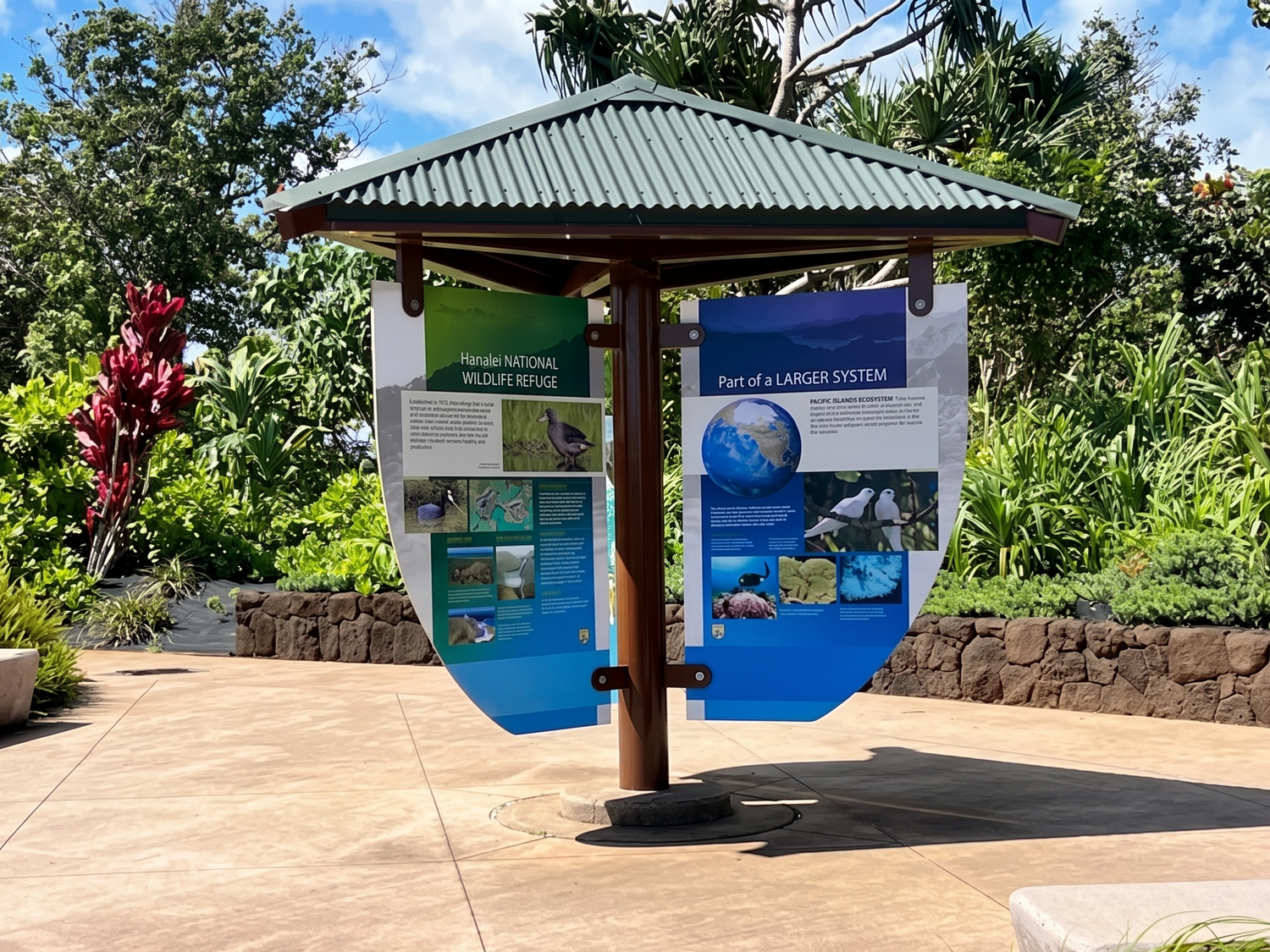 An outdoor educational sign stand with two large hanging panels displaying marine life and ecosystem information, situated on a paved walkway with lush green garden foliage in the background.